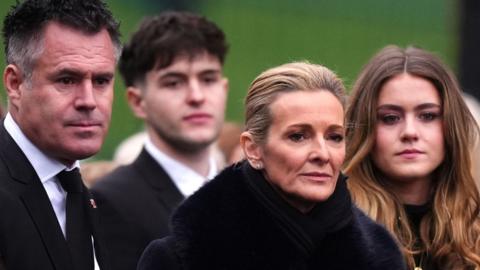 Sports presenter Gabby Logan dressed in black, flanked by her husband and daughter
