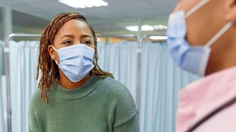 A stock image of a female patient, wearing a mask, talking to a male doctor. Behind them is a privacy screen. 