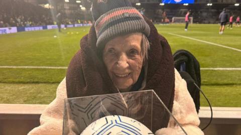 Joan, an elderly woman wearing a woolly hat and with a blanket wrapped around her neck, smiles in the direction of the camera. She is sat in front of a football pitch, where a few players can be seen dotted around. 