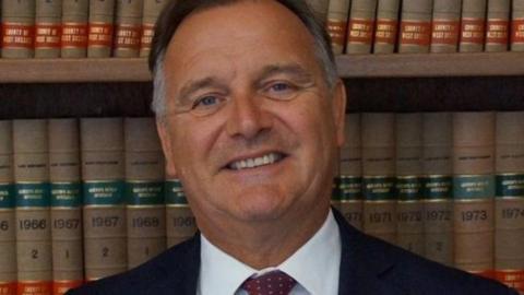 A close up head and shoulders photo of Paul Marshall with a bookshelf behind him. Cllr Marshall is smiling. He has greying short dark hair and wears a black suit, a white shirt and a red tie with small white polkadots.