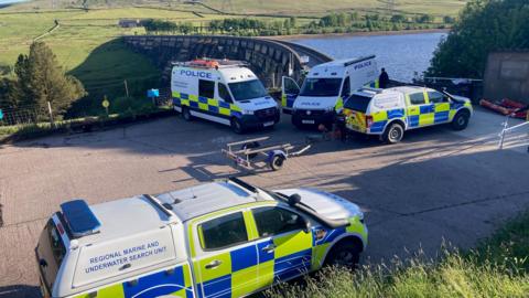 Four police vans, one marked as a marine and underwater search unit, parked outside a reservoir