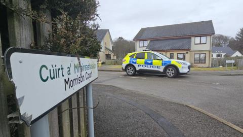 A police car parked in Morrison Court. The SUV is white white blue and yellow markings. It is parked outside a semi-detached house. There is a sign in the foreground that says "Morrison Court" in English and Gaelic.