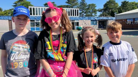 Four children in brightly coloured clothing stand in a school playground and smile at the camera