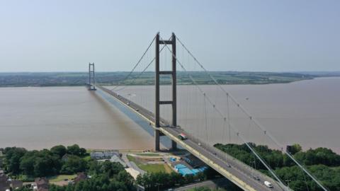 An aerial shot of a large bridge spanning a wide brown river, with tall towers and cables holding it up. Cars are travelling across the bridge. There are houses, trees and a small construction site near the bridge's base in the foreground of the photo. 