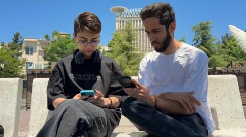 A woman and man sit next to each other on a stone bench looking at their mobile phones in Tehran, Iran
