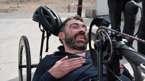 A man lies in a handcycle, with a helmet resting behind his head and team members nearby.