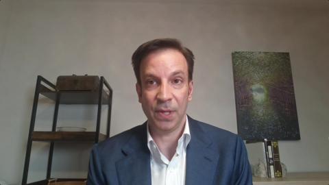 Erik Slavin wearing a white shirt and blue blazer, a grey wall in the background with dark metal-framed shelves with objects on display to his right, and a textured, greenish relief object on a thin stand to his left.