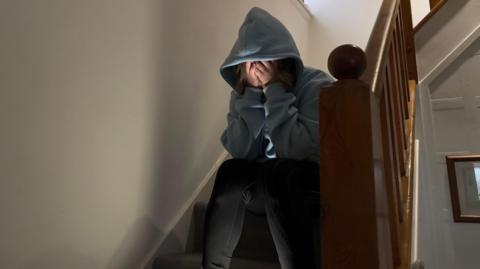 An unidentified woman sitting on a staircase in a house. She is wearing a blue hoodie and black jeans and her face is obscured as she has her head in her hands.