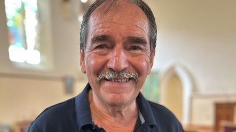 Graham Morgan smiles at the camera. He has a moustache and wears a dark blue polo top. Only head and shoulders in view, behind him the building is blurred but looks to be the inside of a whitewashed church.