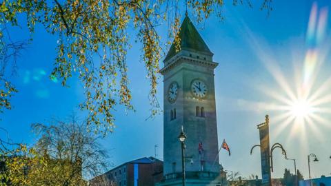 Bright blue sky with sun shining next to a clock tower with two Union Jack flags just beneath. Tree branches with light green leaves in the foreground. 