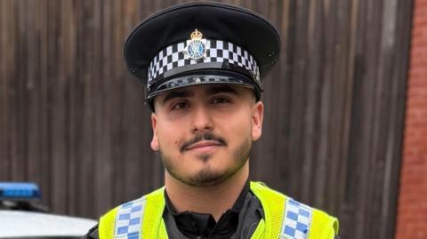 PC Abreu looks at the camera and is wearing a police hat. He is wearing a hi-vis jacket in front of a police car and wooden fence.