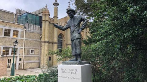 The statue of Max Miller in the Royal Pavilion Gardens, Brighton, with the Brighton Dome and a slightly bent lamppost in the background.