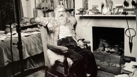 The sculptor Michael Black is pictured reclining in an old armchair in his home. The photo is black and white, and he sits in front of a large stone fireplace. He is wearing baggy black trousers, with a checked shirt over a white top. A bookshelf can be seen along with a surface littered with notebooks and other items