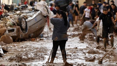 A woman wipes her face as she stands in mud with people and an upturned car in the background