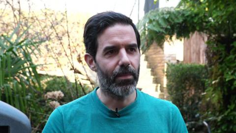 A man stands in a garden wearing a blue T‑shirt with a small microphone clipped to it. Behind him are bushes and trees.