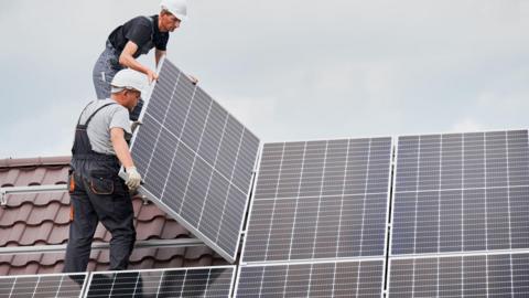 Two men are standing on a roof fitting solar panels
