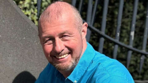 Justin Bellhouse is wearing a light blue, buttoned up shirt. He has thinning hair and a goatee beard. He is smiling.