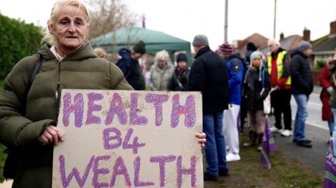 Tracy Lavery stands with other workers on a pavement. She is wearing a padded khaki coat and carries a cardboard sign saying health before wealth.