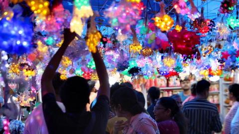 Many colourful lights in the shape of flowers are hanging overhead, with people walking underneath them. A man, on the left, is reaching up with both hands to the ceiling