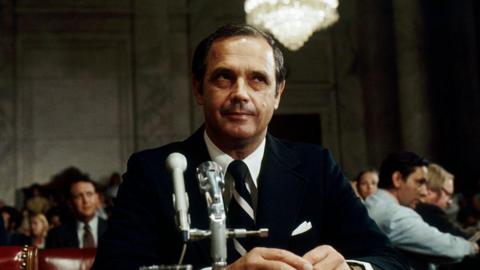 Alexander Butterfield sits before the Senate Watergate Committee wearing a black suit, with a microphone in front of him.