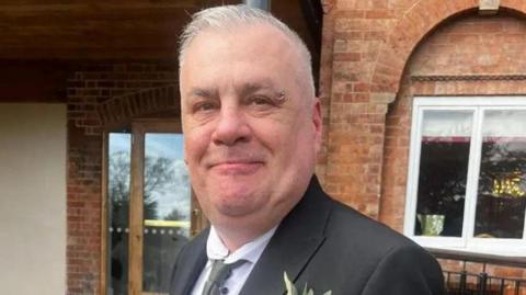 A man with short cropped grey hair and a ring piercing on the end of his left eyebrow. He is wearing a black blazer over a white shirt and a grey tie. There is a flower in the right breast pocket. He is standing in front of a red-brick building and smiling.