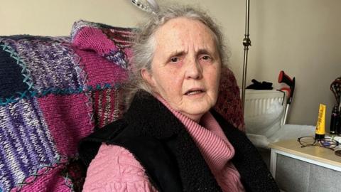 Karen Mitchell, an elderly woman in a pink jumper, is sitting on a chair with a patchwork woollen blanket over it