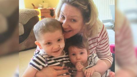 A women is cuddling her two children and smiling at the camera. One of the children has a feeding tube in her nose.