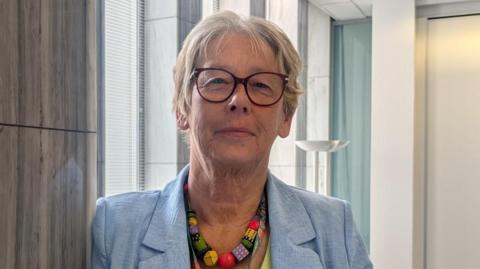 Jane Ashworth wears a light blue jacket, with a bold colourful necklace. She is wearing glasses and smiling at the camera. 