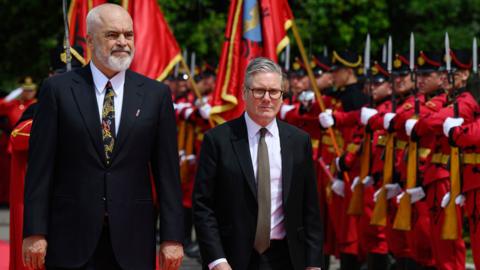 UK PM Keir Starmer is pictured standing next to Albanian PM  Edi Ramat. Behind them, people wearing red military attire stand in a formation; some hold guns, others hold up flags. 