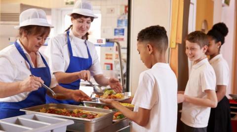 Two ladies, dressed in white shirts, blue aprons and white hats, serve dinners to three pupils, dressed in white polo shirts, in a school. 