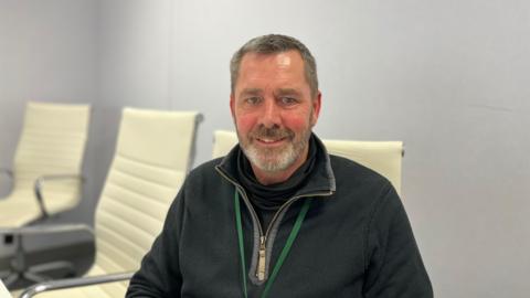 Deputy Marc Leadbeater seated on a cream chair. He has dark grey hair and a grey and brown beard. His cheeks are rosy and he has blue eyes. He is wearing a blue quarter-zip and a green lanyard. 