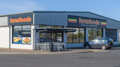 A farm foods store with a metal corrugated roof. A blue car is parked outside in the carpark alongside some shopping trolleys 