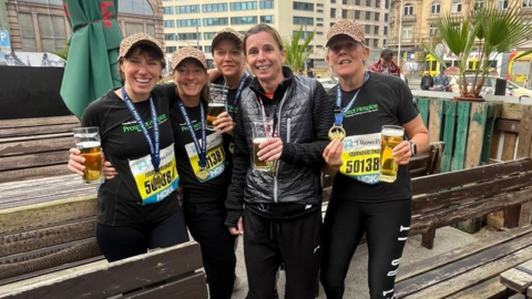 The five women, all smiling to camera, in an outside bar holding pint glasses, and still wearing their black marathon gear, including race bibs and medals