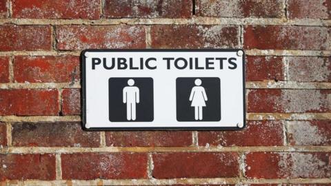 A public toilet sign screwed to an exterior brick wall. There are symbols indicating male and female cubicles.