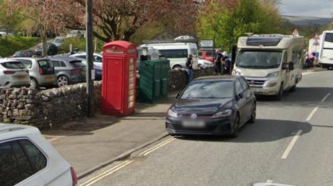 A traditional red phone box next to a busy car park with cars, vans, a motorhome and a coach.