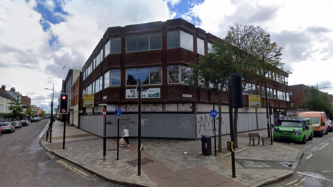 The brown and glass building stands on the corner of two streets. It is boarded up on the lower level and has graffiti on it. A tree can be seen in the paving on the right of the centre with vehicles parked on one side street and traffic lights on the left side.