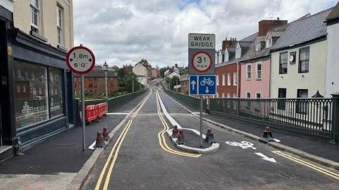 A narrow bridge with road signs including a 1.8-meter (6 feet) height restriction on the left, a central warning for a weak bridge with a 3-ton weight limit, and a blue sign allowing bicycles to proceed straight. The road has yellow lane markings and bollards guiding traffic. Red barriers are visible on the left sidewalk, and buildings line both sides of the bridge.