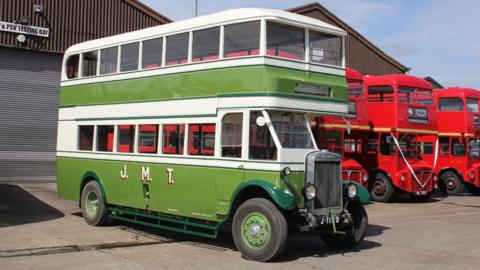 A green Jersey TV 1 bus on a sunny day. There are other red busses parked in a row beside it.