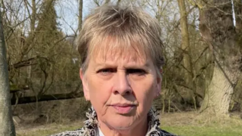 Woman, with blonde hair, wearing a black and white jacket and white shirt, looking expressionless, with a trees and grass behind her.