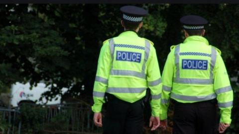 Two police Scotland officers viewed from behind as they look into a wooded area.