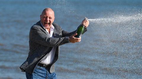 John Lingard wearing a grey suit jacket with a white shirt and jeans. He is stood on a beach and is smiling at the camera with a bottle of champagne. 