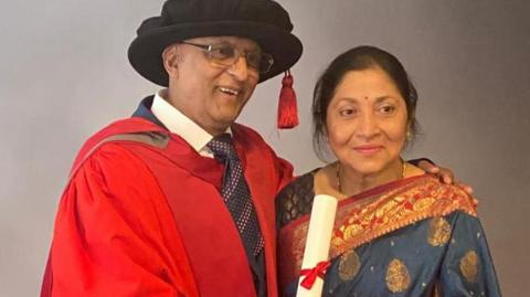 Needyanand Raya and his wife Lacksoomi pose against a grey background in a graduation photo. He is wearing a red graduation robe and hat, while holding a paper diploma. His wife is pictured in a patterned blue and red saree.