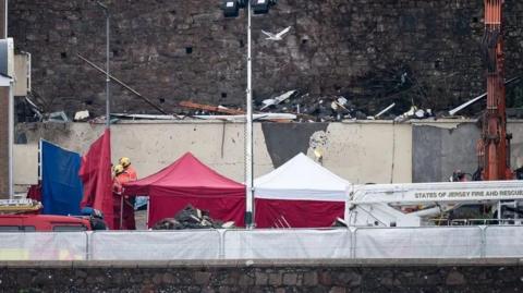 Rubble and aftermath of the fire next to pop-up tents for emergency workers, two of which, wearing safety helmets, are talking with each other 