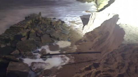 An eroded sand dune pictured at night time. The sea is visible and what appears to be a metal rod.