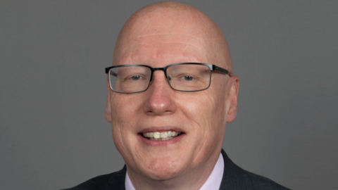 A man with a clean-shaven head and metal-framed glasses smiles at the camera against a grey background in a corporate headshot.
