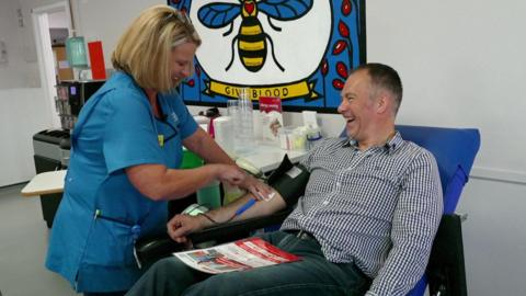 Roger Johnson, who has short hair and is sitting on a blue chair in a medical room, is giving blood. A nurse with blonde bobbed hair and wearing a light blue uniform is collecting blood from Roger's arm. They are both smiling.