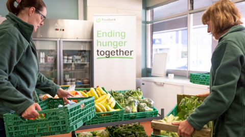 Two women dressed in Trussell Trust green hold pallets of vegetables in front of a sign that says 'ending hunger together'