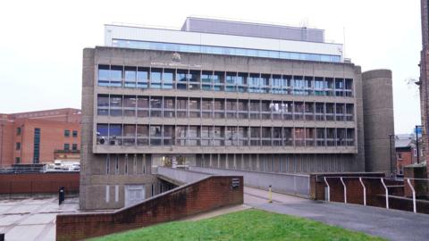 A grey brutalist building, pictured from the other side of a footbridge which leads to it. The building has lots of windows. It's a cloudy day.