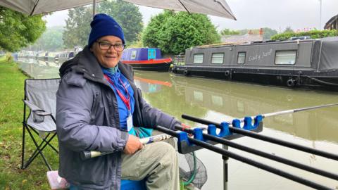 A woman in a blue woolly hat, blue jumper and navy coat sits on a bright blue box holding a fishing rod by the canal. 