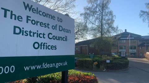 A green and white sign for the Forest of Dean District Council stands under blue skies in front of the council offices.